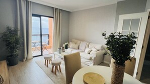 Una sala de estar moderna con sofá, mesa de centro y sillas. Hay un balcón con vista al mar.