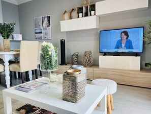 A modern living room with a television displaying a news program, a white coffee table with magazines and decorative items, a dining area with a vase of flowers, and a bookshelf with various decorative objects.