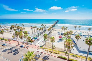 A coastal cityscape with a long pier extending into the ocean, palm trees lining the beachfront, and a clear blue sky. - California Craftsman Beach Bungalow in Oceanside! (Oceanside)