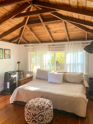A bedroom with a wooden ceiling, a bed with white bedding, a patterned ottoman, and a desk with a lamp.