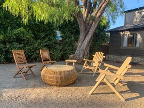 An outdoor seating area with wooden chairs and a round table, surrounded by dense foliage and a large tree.
