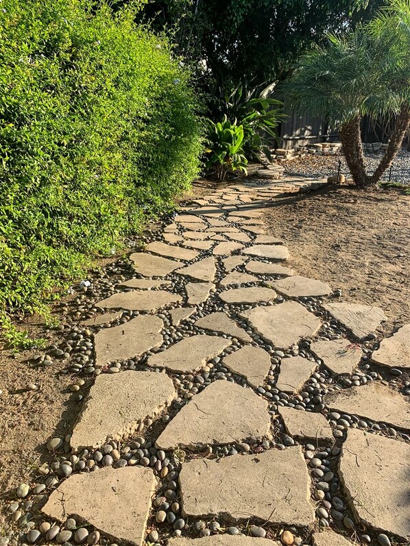 A stone pathway with irregularly shaped stones and small pebbles, surrounded by lush greenery and trees. - California Craftsman Beach Bungalow in Oceanside! (Oceanside)