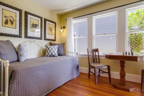 A bedroom with a bed, a chair, a table with chess pieces, and framed pictures on the wall.