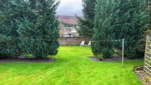 A well-maintained backyard with a grassy lawn, two large evergreen trees, a stone wall, and a view of a hill in the background.