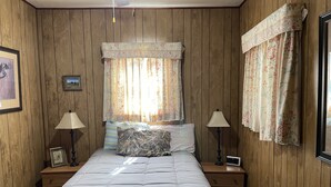 Een slaapkamer met houten lambrisering, een bed met een grijze dekbedovertrek, twee nachtkastjes met lampen, een nachtkastje met een klok, en een raam met bloemen gordijnen.