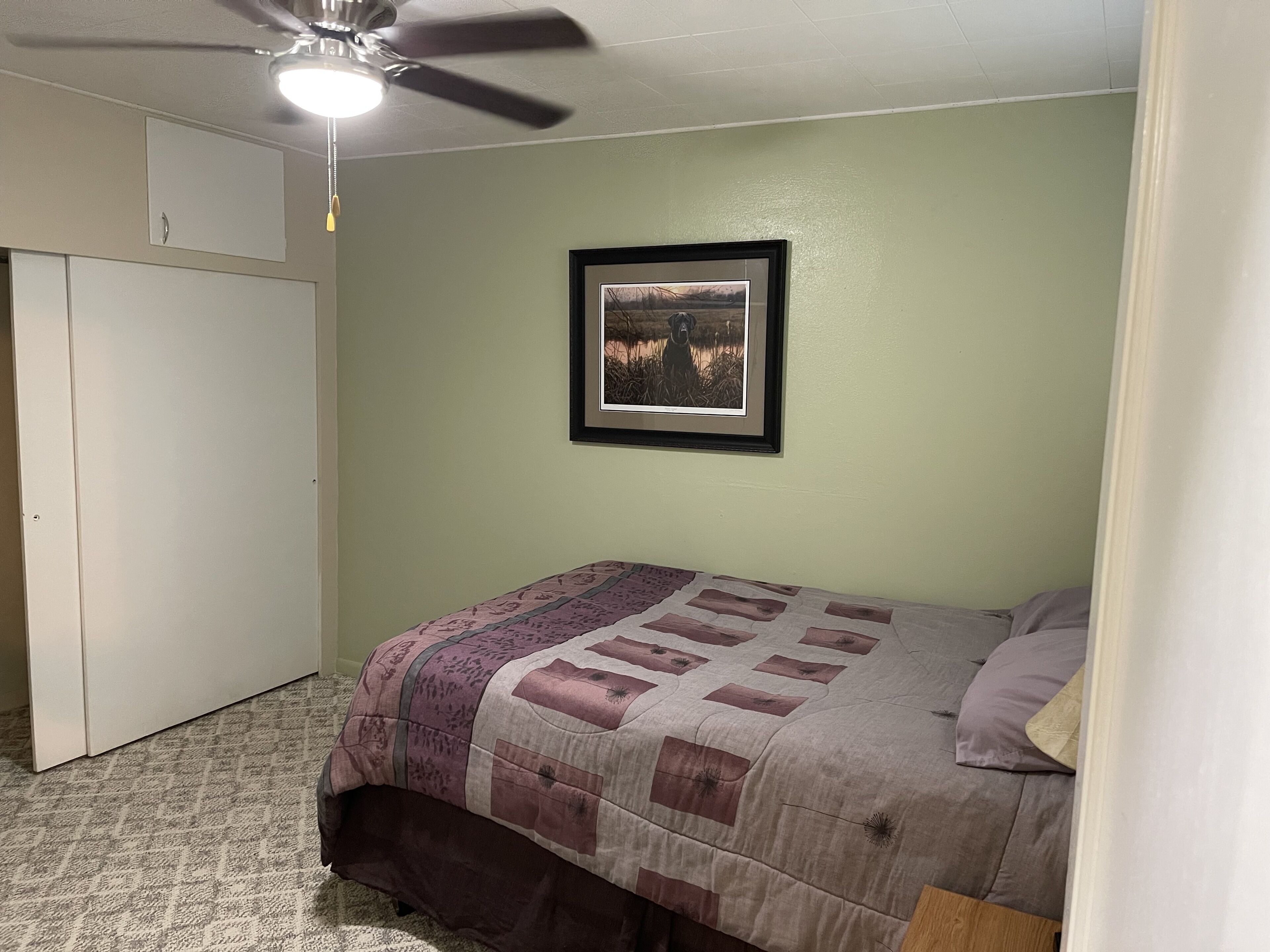 Een slaapkamer met een bed, een plafondventilator, een lijst met een afbeelding aan de muur en een kledingkast.
