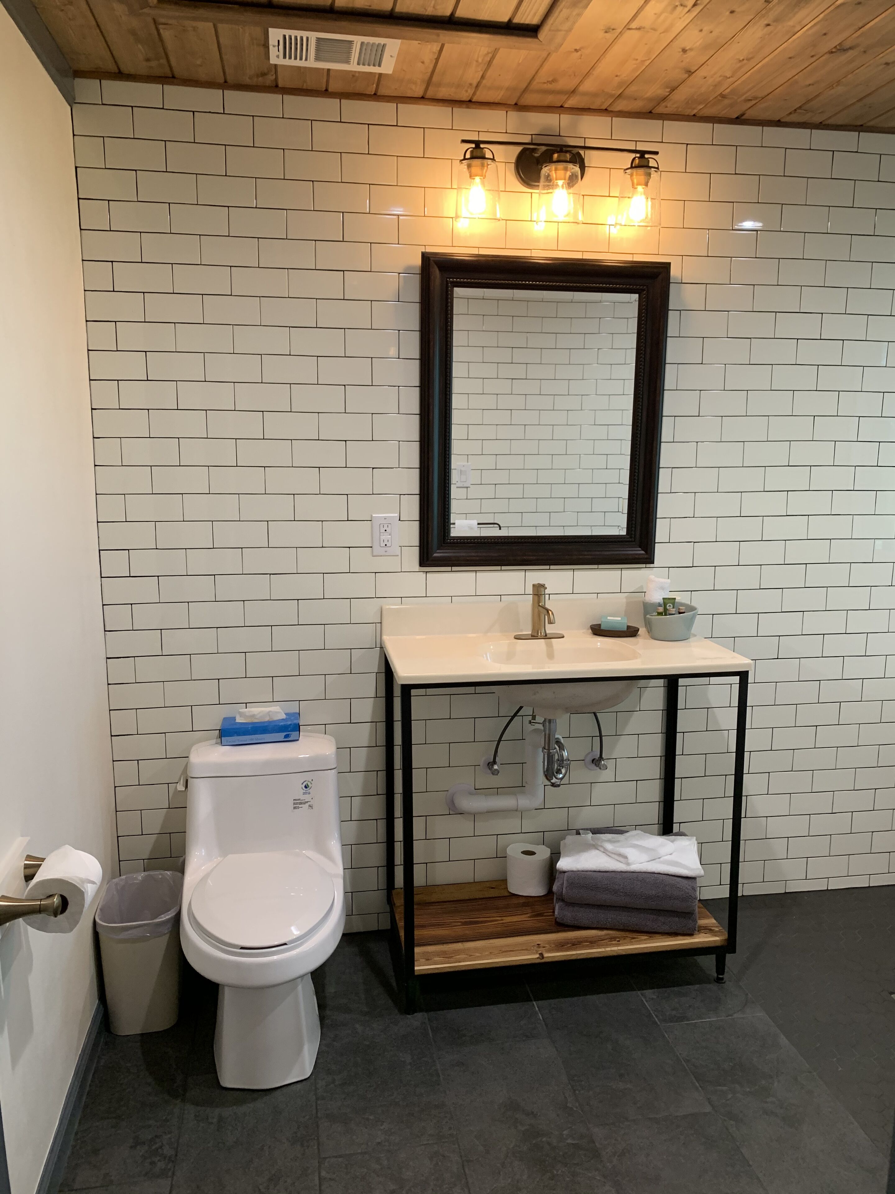 Un bagno con parete rivestita di piastrelle bianche, soffitto in legno, water bianco, lavabo con cornice nera e specchio sopra il lavabo.