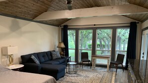 A bedroom with a bed, a sofa, a small table, and chairs. There is a large window with curtains, and a view of trees outside. - Salado Cottage Retreat near Downtown (Salado)