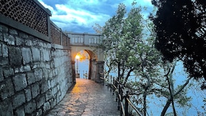 Ein Steinweg führt zu einer Brücke über dem Wasser, mit einem Geländer auf einer Seite und einer Lampe am Ende.