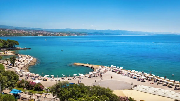 Ein Küstenstrand mit Liegestühlen und Sonnenschirmen, ein klares blaues Meer und eine Ansicht der Stadt in der Ferne.