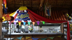 Een restaurant met rieten dak, een toonbank met een menu en kleurrijke decoraties, waaronder een spandoek met de tekst 'The Greatest Showman'.