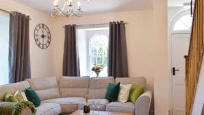 A living room with a sectional sofa, a wooden coffee table, a chandelier, a clock, and a window with curtains.