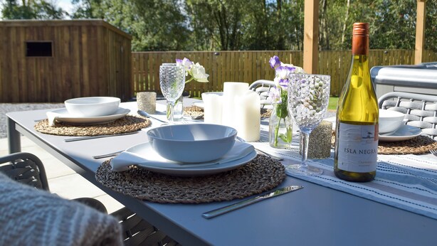 An outdoor dining setup with a table set for two, featuring a bottle of Isla Negra wine, white plates, bowls, and a woven placemat.