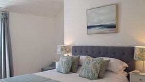 A neatly made bed with a grey headboard, white bedding, and patterned pillows. There is a bedside table with a lamp on each side of the bed. A painting hangs above the bed, and a curtain is partially visible on the left.