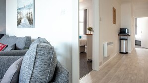 A modern living room with a grey sofa, a painting on the wall, and a hallway leading to a bedroom.