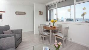 A modern living room with a grey sofa, a dining table with chairs, a vase with flowers, and a large window with a city view.
