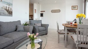 A modern living room with a grey sofa, a glass coffee table with a vase of flowers, and a dining area with a wooden table and chairs.