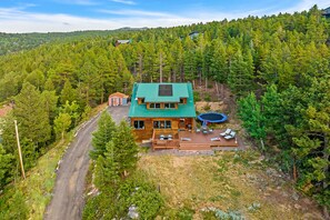 A cabin with a green roof, surrounded by dense forest, featuring a deck with outdoor seating and a covered area. - Moss Rocks Cabin~starlink~AC~solar powered ~Ev charger~fire pit ~views  (Nederland)