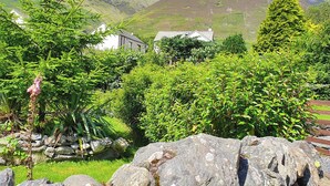 Paisaje rural con muro de piedra, abundante vegetación y una casa de fachada blanca y techo oscuro.