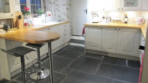Una cocina con armarios blancos, encimera de madera, un taburete de bar y suelo de piedra.