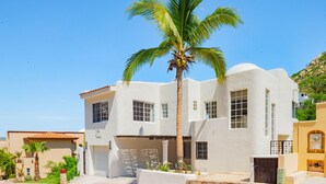 Un edificio de dos pisos con fachada blanca, una palmera frente a él y un cielo azul despejado.