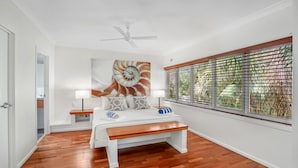Un dormitorio con piso de madera, una cama con banqueta, un ventilador de techo y una ventana con persianas.