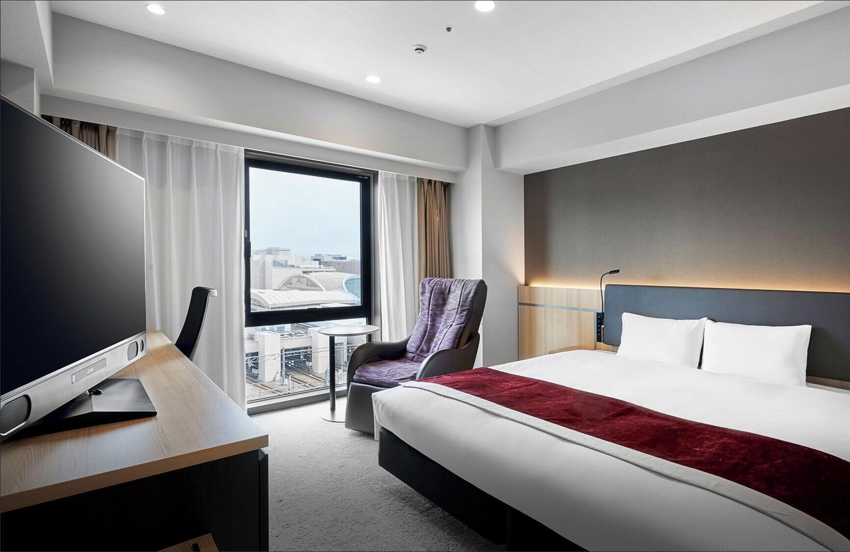 Quarto de hotel com uma cama grande, uma escrivaninha com TV de tela plana, uma cadeira, uma mesinha e vista da cidade por grandes janelas.