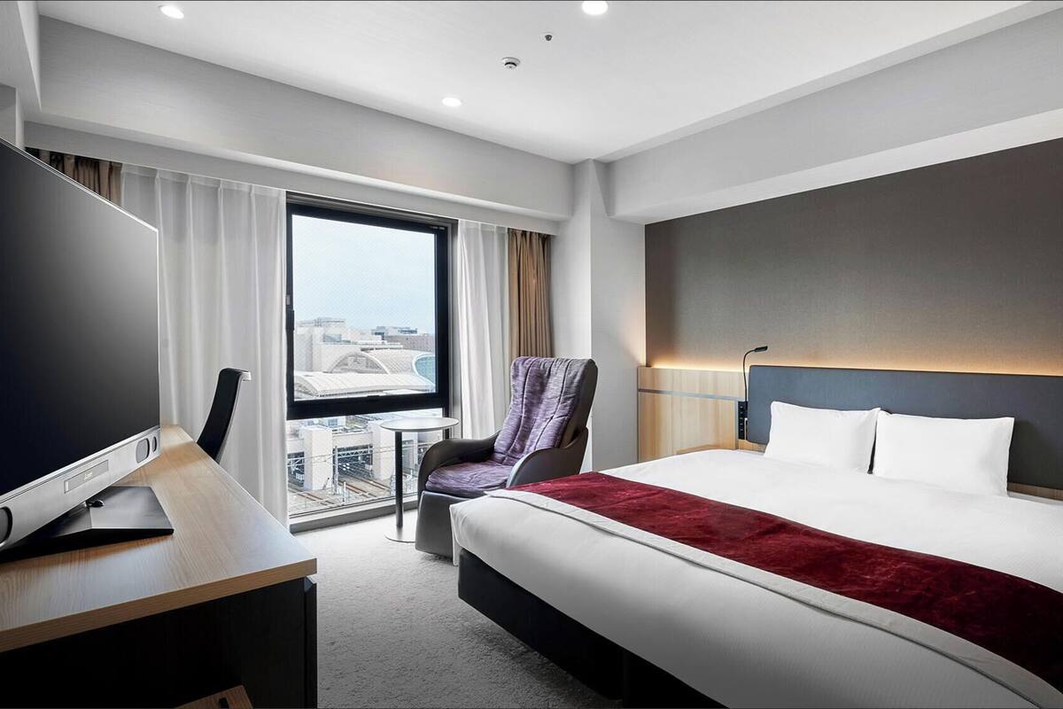 A hotel room with a large bed, a desk with a flat-screen TV, a chair, a small table, and a view of the city through large windows.