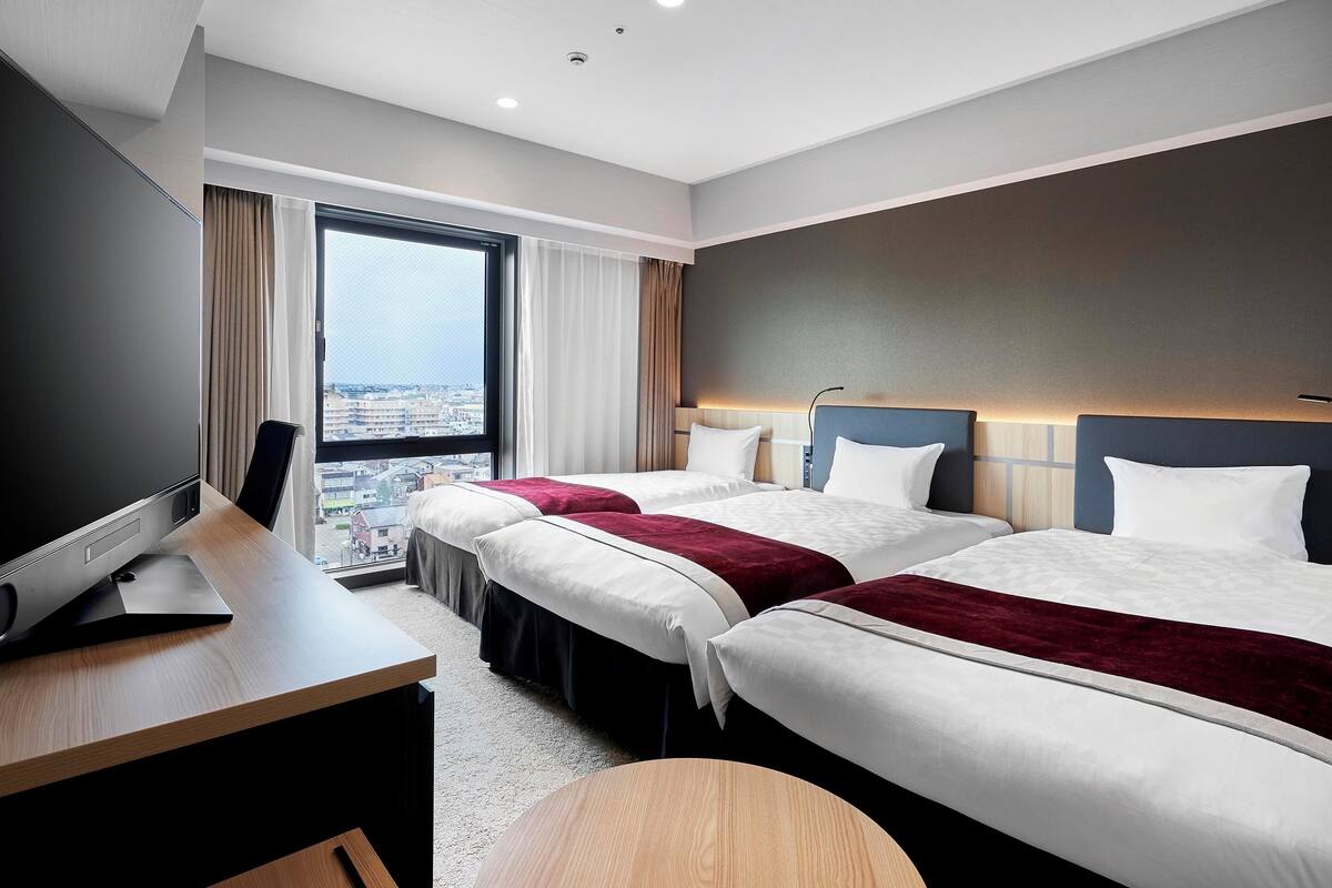 Quarto de hotel com duas camas, uma escrivaninha, televisão e uma janela grande com vista para a cidade.
