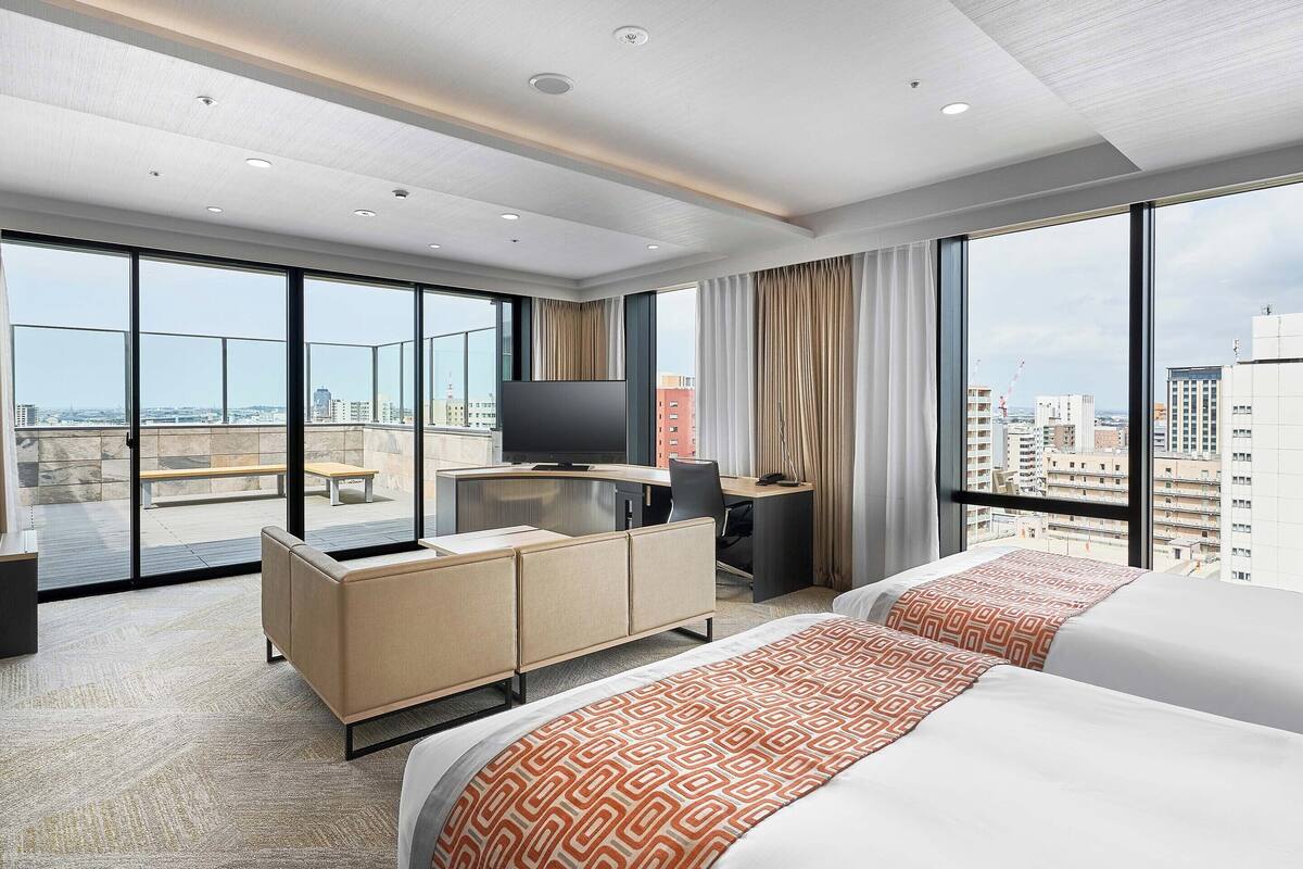 Quarto de hotel com uma janela grande, uma escrivaninha, uma televisão e duas camas.
