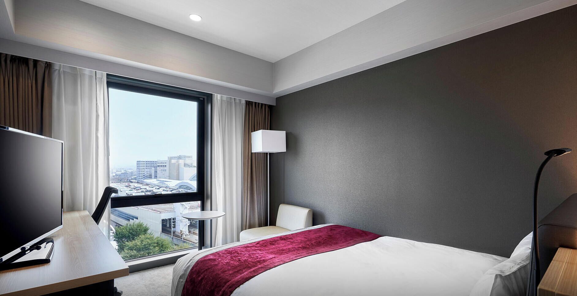 Quarto de hotel com cama, escrivaninha, cadeira, abajur e vista para a cidade.