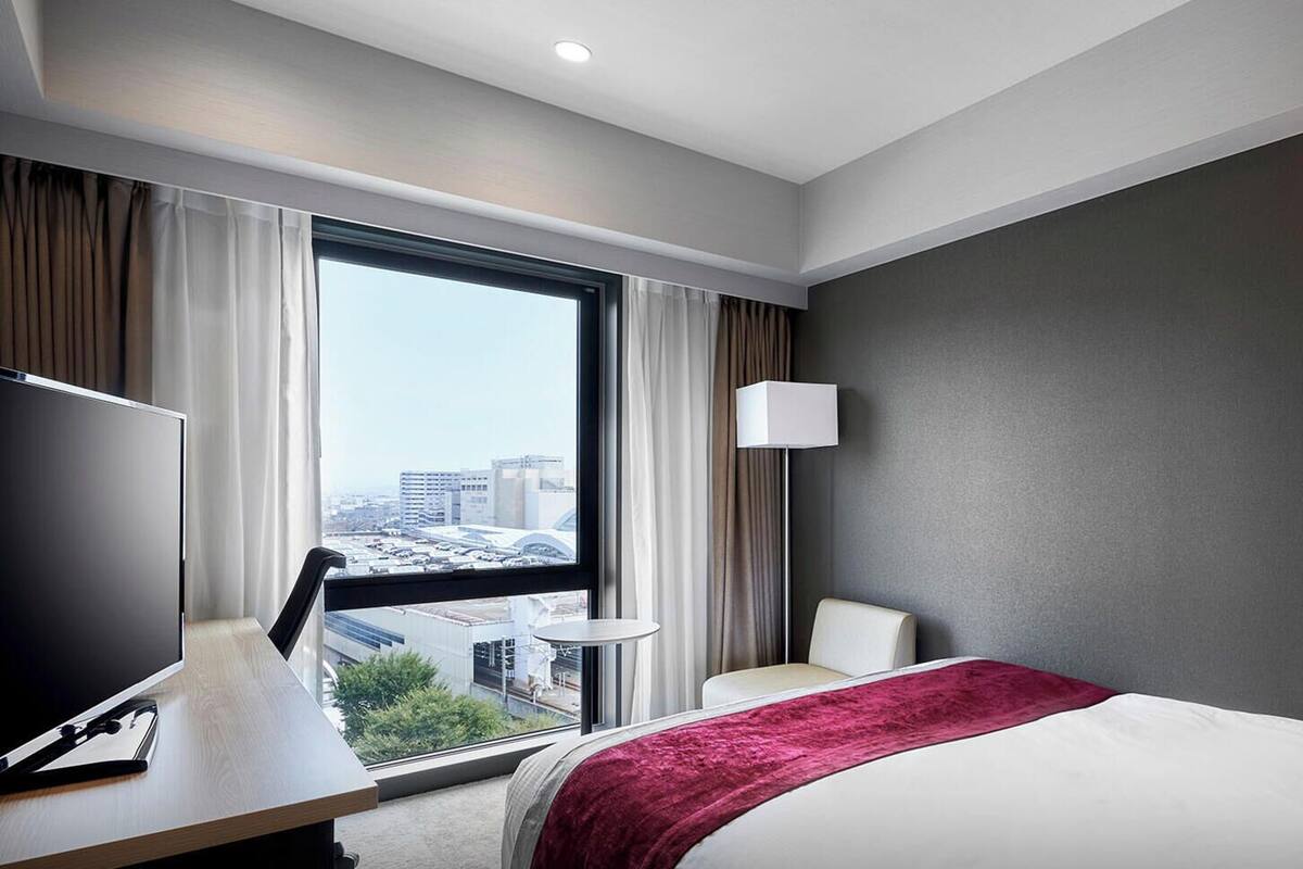 A hotel room with a bed, a desk, a chair, a lamp, and a view of the city.