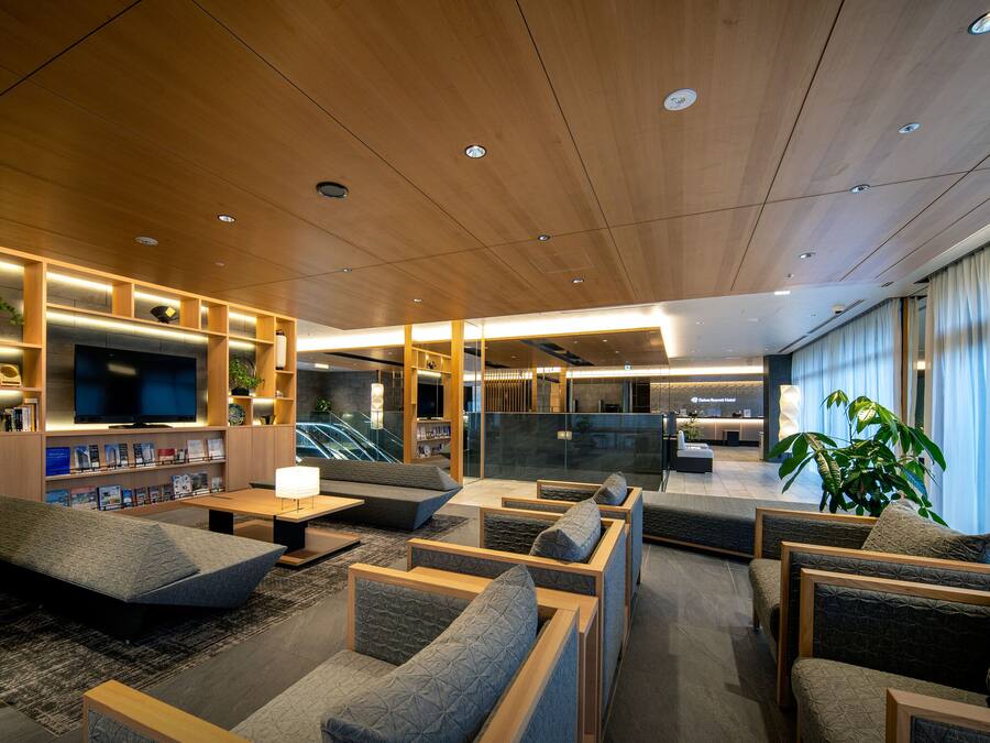 A modern hotel lobby with wooden panel ceilings, a flat-screen TV, seating areas, and a staircase leading to an upper floor.