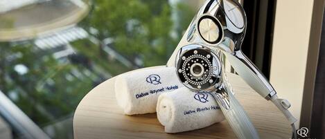 A towel with the Daiwa Roynet Hotels logo, a showerhead, and a glass of water on a wooden surface by a window.