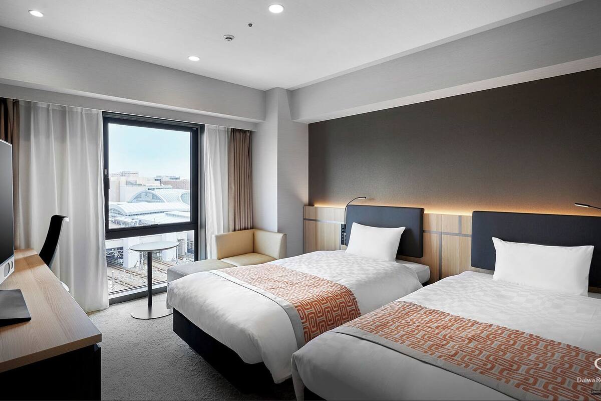 A hotel room with two beds, a desk, a TV, and a view of a cityscape.