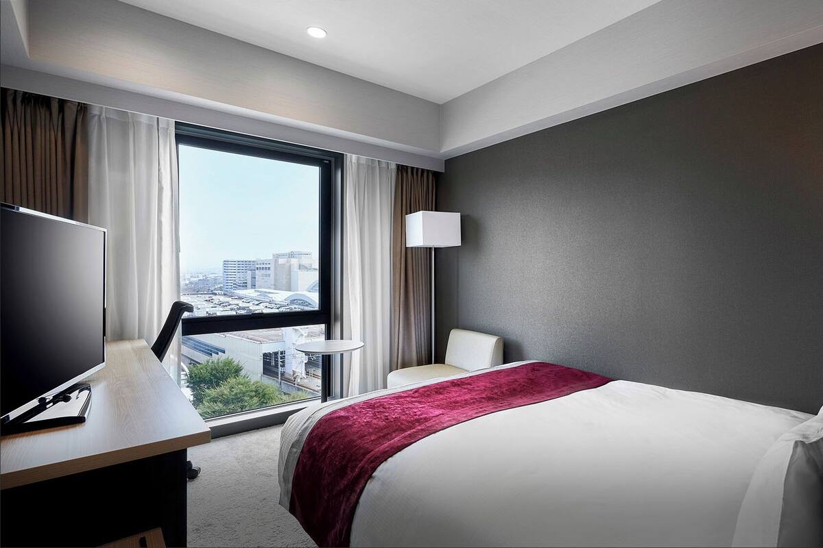 A hotel room with a bed, a desk with a TV, a lamp, and a view of the city through large windows.