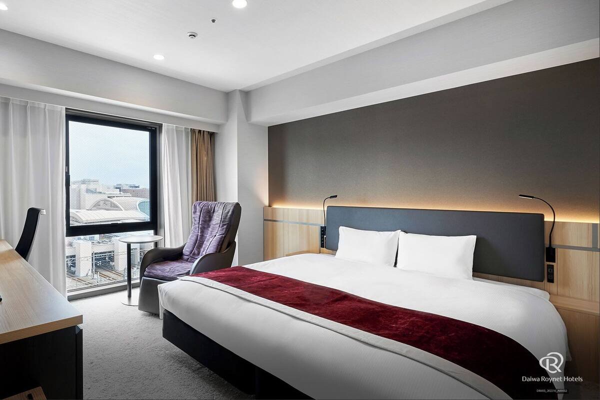 A hotel room with a large bed, a desk with a flat-screen TV, a chair, and a view of the city through a large window.