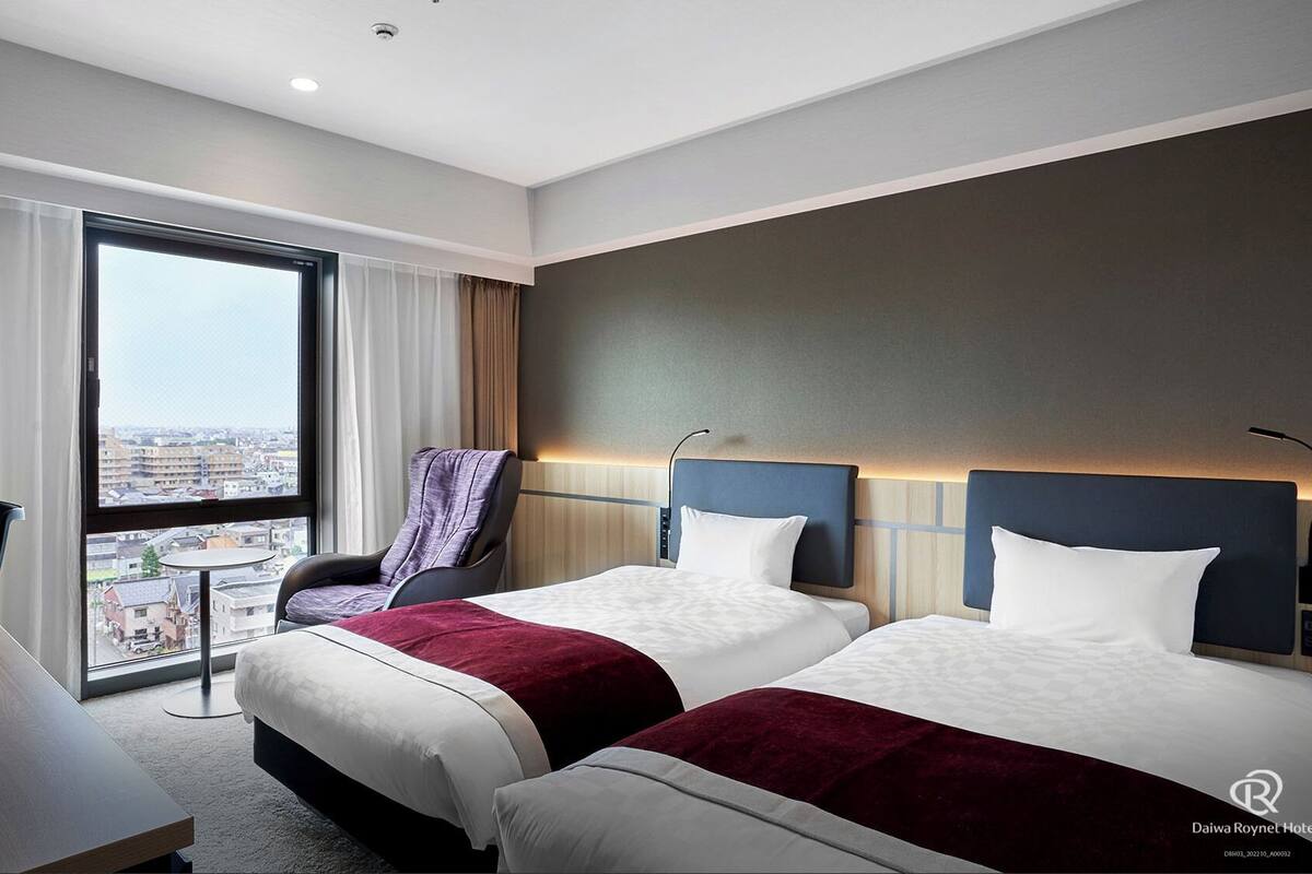 A hotel room with two beds, a desk, a chair, and a large window offering a city view.