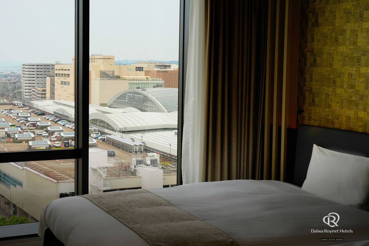 A hotel room with a large window offering a view of a cityscape, including a train station and various buildings.