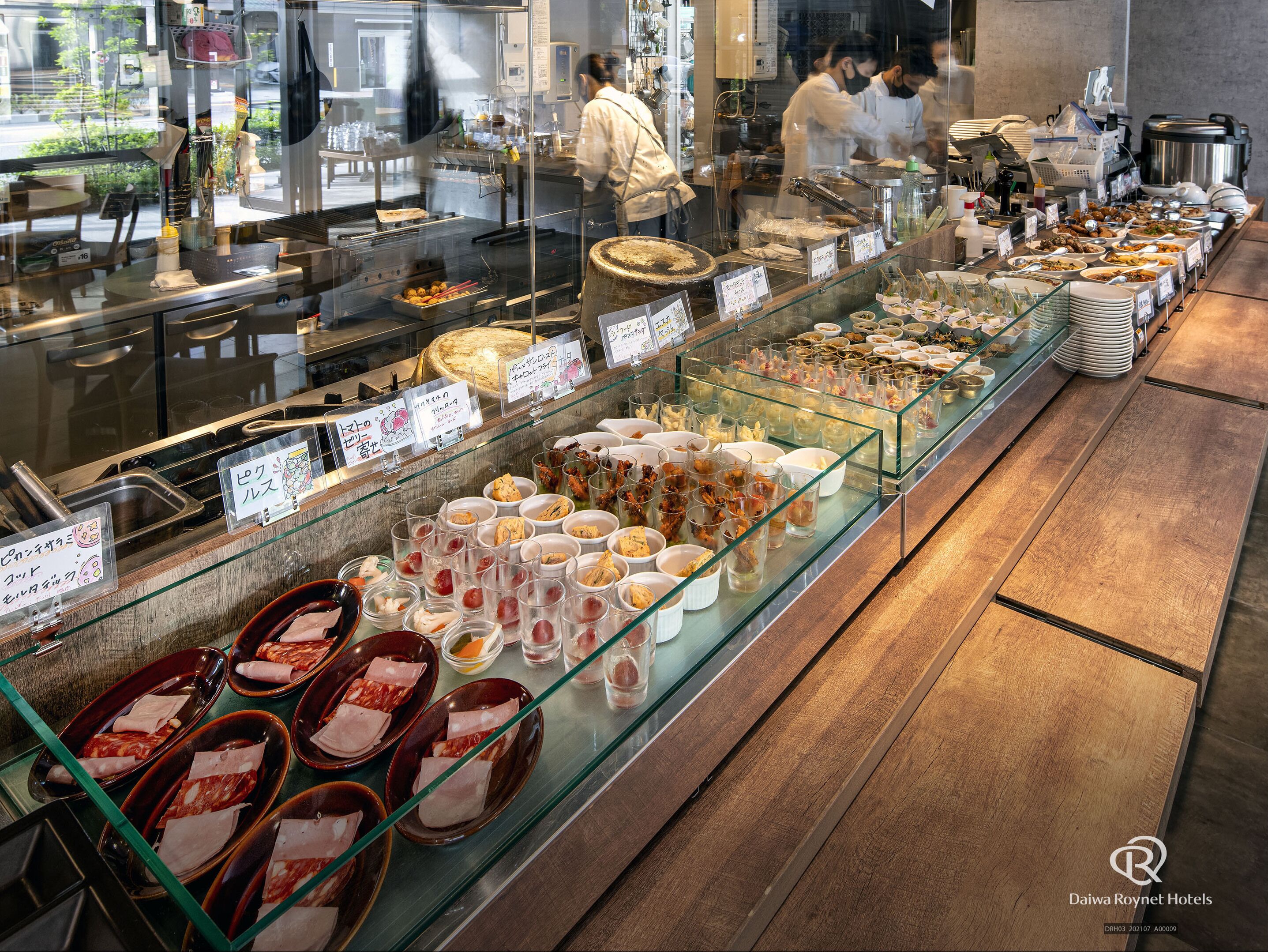 A buffet with various dishes and drinks, a kitchen area with staff, and a wooden floor.