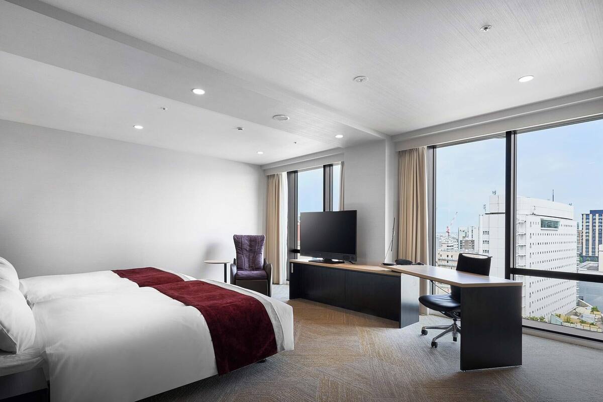 Quarto de hotel moderno com uma cama grande, uma escrivaninha, uma cadeira, uma televisão e vista para a cidade.