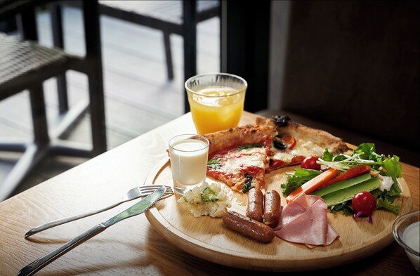 A breakfast plate with ham, sausages, a slice of pizza, a glass of orange juice, and a cup of milk on a wooden table.