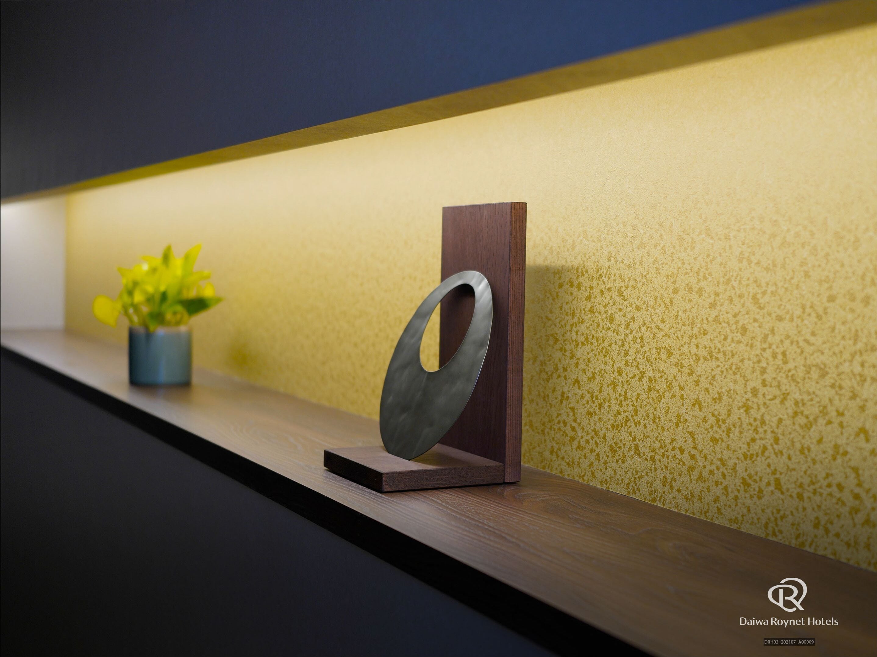 A wooden shelf with a metal ring, a small potted plant, and a textured yellow wall.