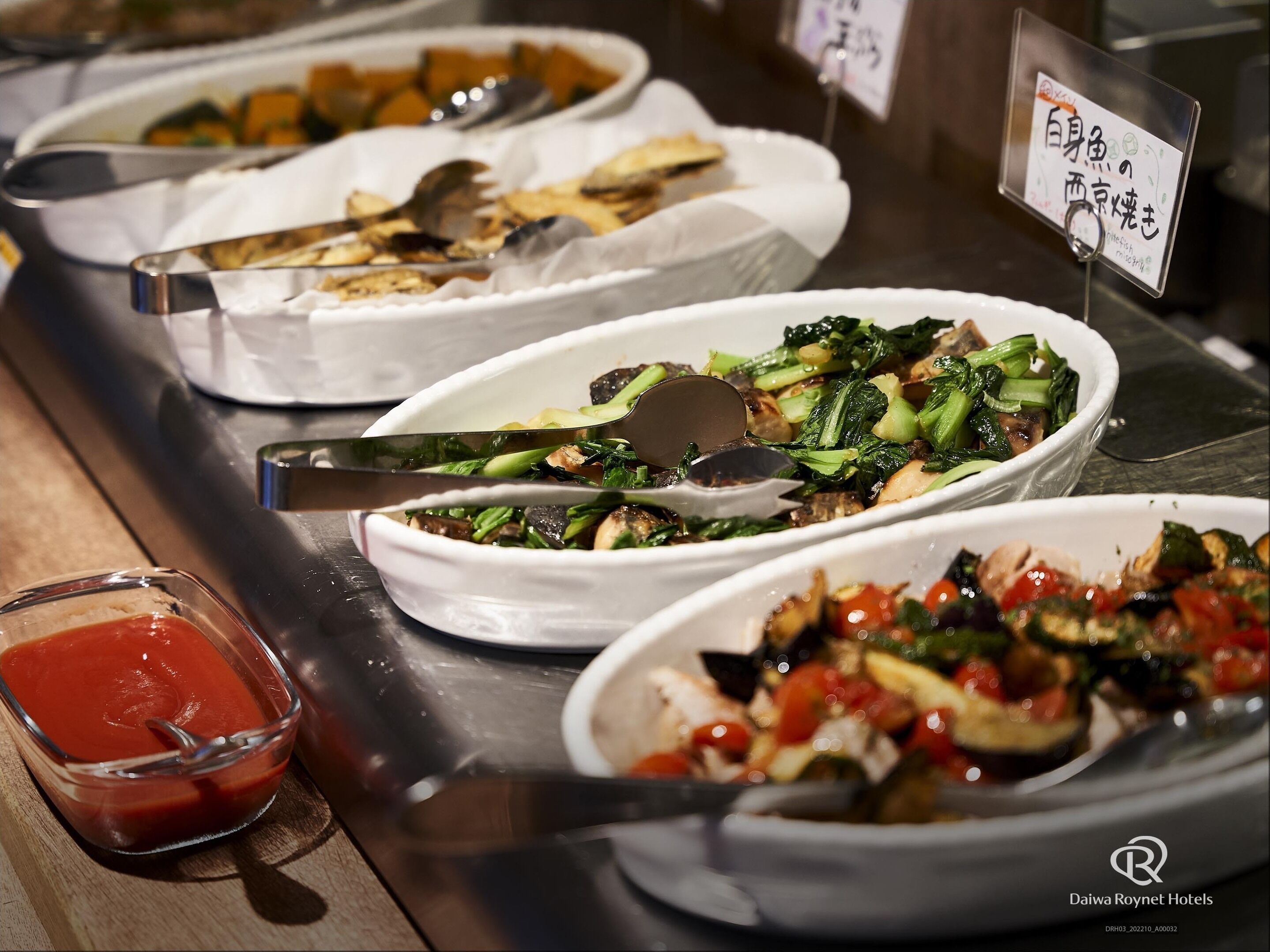 A buffet with various dishes, including vegetables and a bowl of sauce.