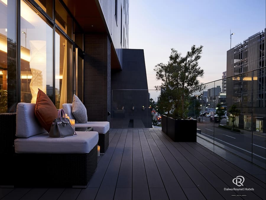 Un área de estar al aire libre con muebles de mimbre y cojines, una planta en maceta y vistas de la ciudad.