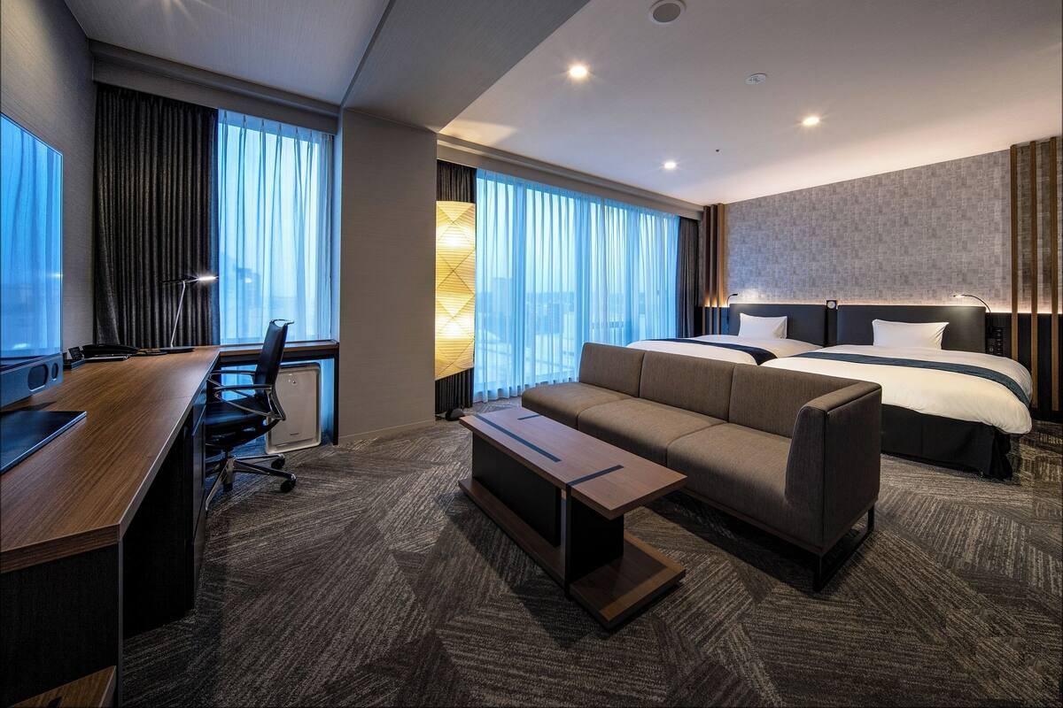 Quarto de hotel moderno com uma cama grande, um sofá, uma escrivaninha e vista para a cidade por grandes janelas.