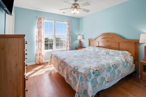 A bedroom with a wooden bed, a nightstand with a lamp, a window with curtains, and a ceiling fan.