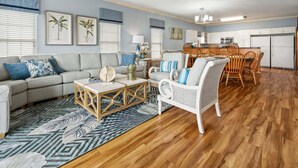 Un salon comprenant un canapé, une table basse et un fauteuil en osier, un coin repas avec des chaises en bois, et une cuisine avec des meubles blancs.