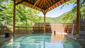 Piscina exterior cubierta con vigas de madera y valla de madera, rodeada de exuberante vegetación y colinas.