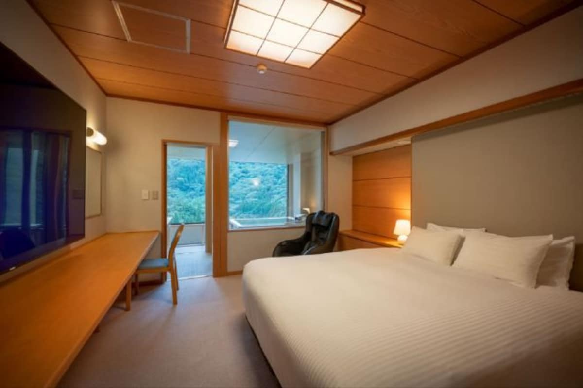 A hotel room with a large bed, a desk, a chair, and a window with a view of greenery.